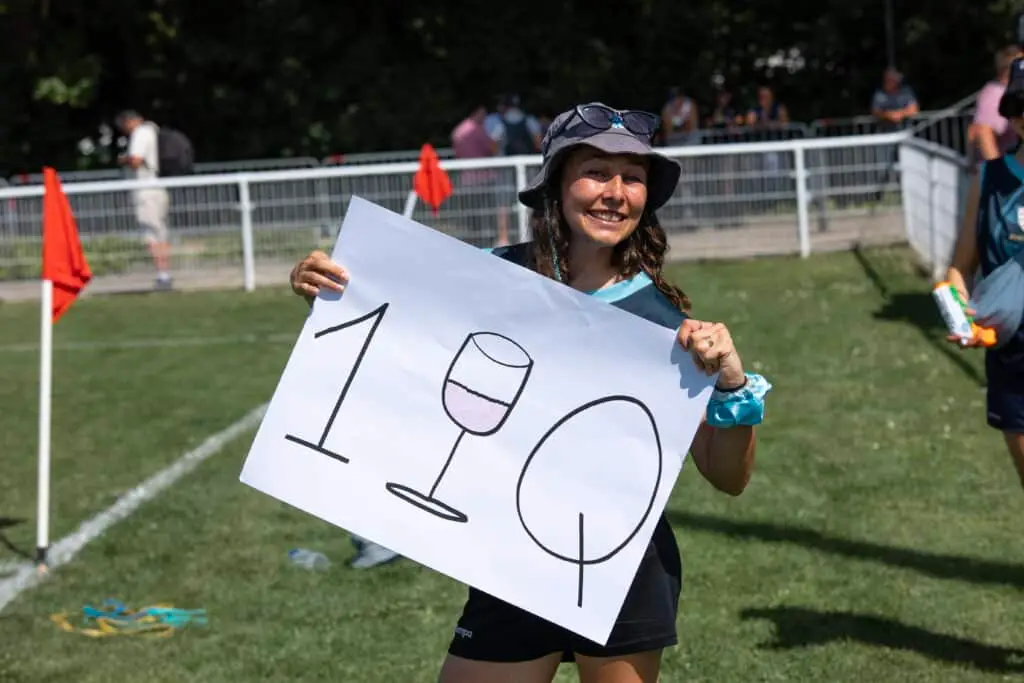 A smiling supporter in a sun hat holds up a hand-drawn sign at the edge of the pitch showing a "1", a wine glass and a rugby ball