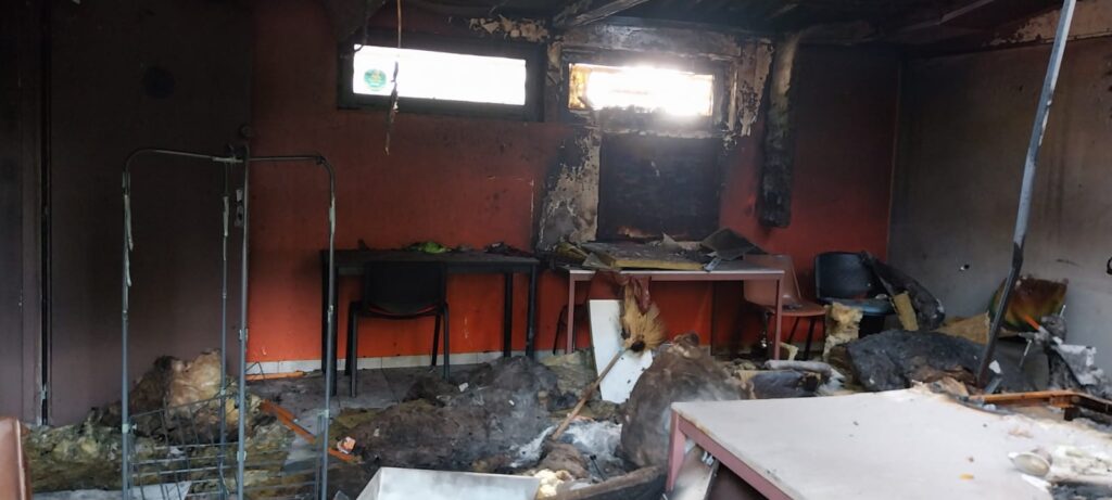 Burnt-out interior of the clubhouse after the fire: blackened walls, insulation collapsed onto the floor, tables and chairs overturned among the debris